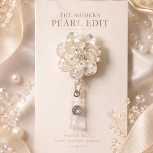 May include: A badge reel featuring a white flower with layered petals and a crystal center, attached to a clear plastic strap with a circular crystal accent. The item is mounted on a cream card labeled THE MODERN PEARL EDIT and BADGE REEL.
