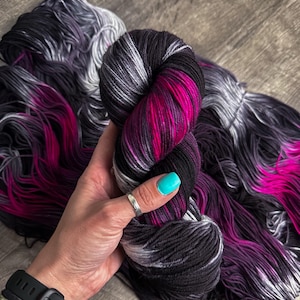 May include: A skein of hand-dyed yarn featuring a variegated colour palette of deep black, charcoal grey, and vibrant magenta pink. The yarn is twisted into a compact bundle, showcasing a blend of dark tones contrasted by bright streaks of pink.