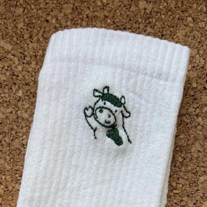 Könnte beinhalten: Weiße gerippte Socke mit einem grün gestickten Cartoon-Kuh-Design. Die Kuh winkt mit einem lächelnden Gesicht. Die Socke besteht aus einem weichen, bequemen Material.