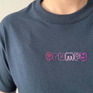 Könnte beinhalten: Ein dunkelblaues T-Shirt mit Rundhalsausschnitt, auf dessen Brust das Wort GRUMPY in einer verspielten, abgerundeten Schriftart gestickt ist. Die Stickerei verwendet einen Farbverlauf aus rosa und violetten Fäden, der sich vom dunklen Stoff abhebt.