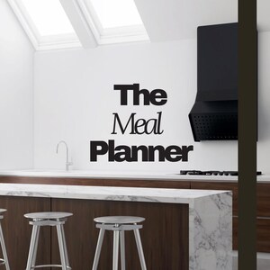 May include: A modern kitchen with a white marble countertop island and three silver stools. The words "The Meal Planner" are displayed on the white wall. A black range hood is above the countertop. Natural light streams in from the skylights.