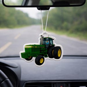 Puede incluir: Un ambientador con forma de tractor verde y amarillo colgado del espejo retrovisor de un coche. El ambientador tiene el texto "4705 BARN & BLOOM". El fondo muestra una carretera y &aacute;rboles borrosos.