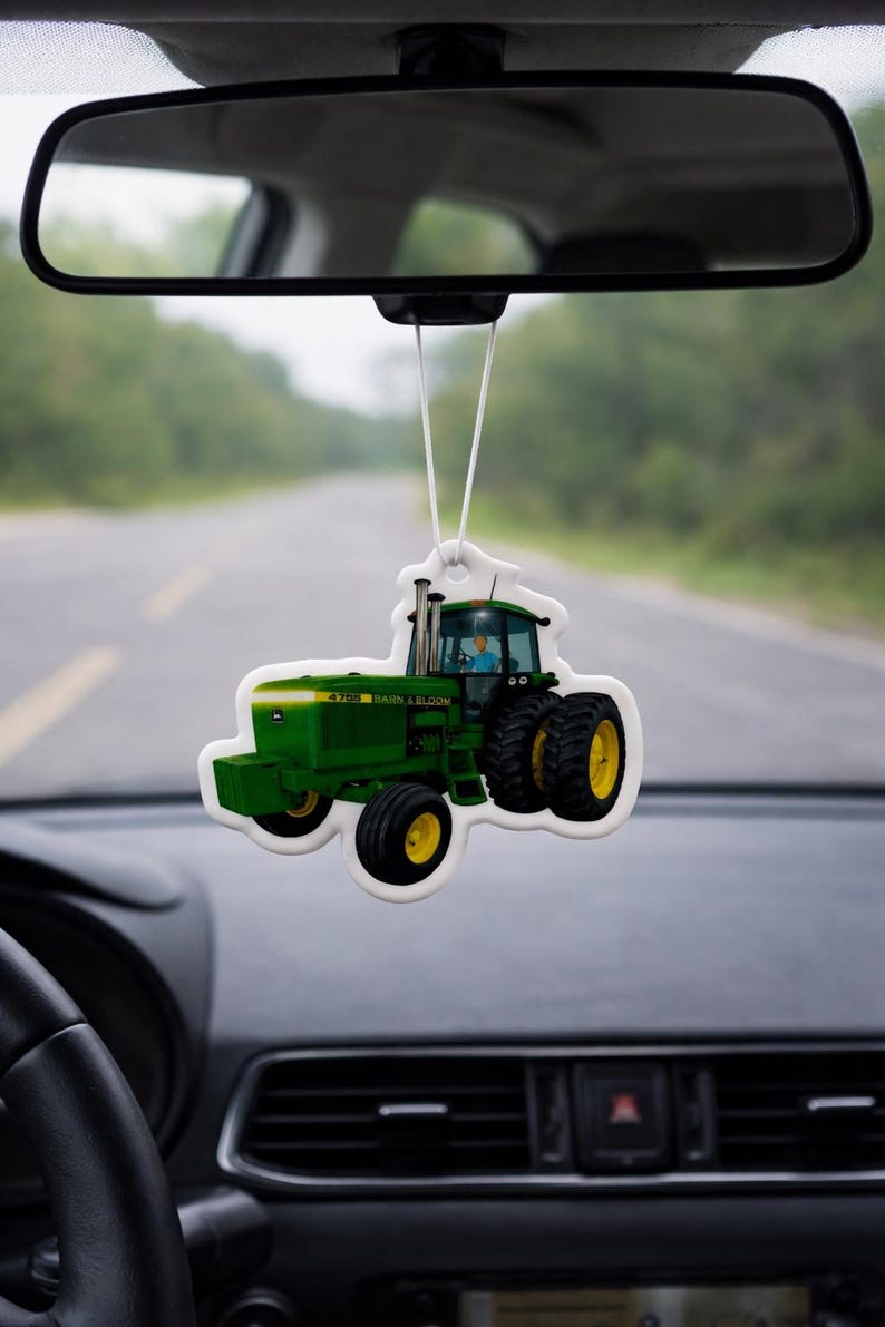 Puede incluir: Un ambientador con forma de tractor verde y amarillo colgado del espejo retrovisor de un coche. El ambientador tiene el texto "4705 BARN & BLOOM". El fondo muestra una carretera y &aacute;rboles borrosos.