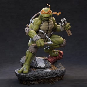 May include: A detailed statue of Michelangelo from the Teenage Mutant Ninja Turtles. The figure is green with an orange mask, holding nunchucks. The statue is posed on a rocky base with a red fire hydrant.