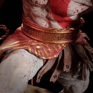 May include: A detailed figurine torso featuring pale skin with red markings and blood splatters. The figure wears a metallic bronze belt with a Greek key pattern, a red fabric wrap with gold trim, and brown leather tassets with dark metal rivets.