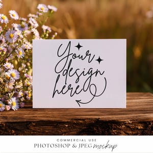 以下が含まれることがあります： 黒い筆記体で「Your design here」と書かれた白いカード。星と矢印のアクセントが付いています。カードは木製の表面にあり、背景にはぼやけた花と葉があります。画像はモックアップです。