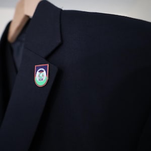 May include: A navy blue blazer with a small, colorful pin attached to the lapel. The pin features a crest design with green, white, and blue elements. The blazer is hanging on a wooden hanger.