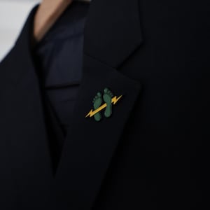 May include: A dark navy blazer with a green and yellow pin. The pin features two green footprints with a yellow lightning bolt. The blazer is a solid color and appears to be made of a smooth fabric.