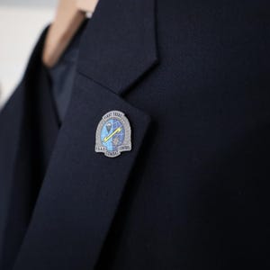 Może przedstawiać: Zbliżenie granatowej marynarki z przypinką w kolorze srebrnym na klapie. Przypinka ma napis "FIRST TROOP U.S.A.F. COMBAT CONTROL". Marynarka wisi na drewnianym wieszaku.