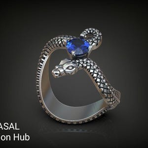 May include: A silver-colored ring in the shape of a snake, with a blue gemstone at the top. The snake's body is textured with a scale pattern. The ring is on a dark background. Text on the bottom left says "RASAL Fashion Hub".