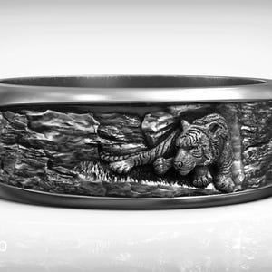 May include: A silver-toned ring with a detailed relief carving of a tiger resting in a naturalistic setting. The ring's band is wide, and the tiger is depicted with intricate fur and facial features. The ring is a piece of jewelry.