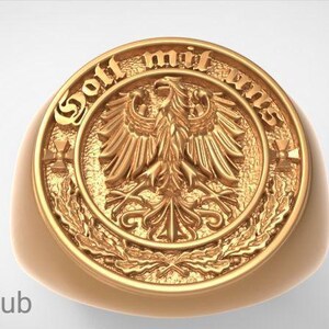 May include: A gold-colored ring with a detailed eagle emblem at the center, surrounded by the words "Gott mit uns". The ring has a rounded shape and a polished finish, with the brand name "RASAL Fashion Hub" in the lower left corner.