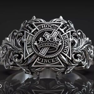 Könnte beinhalten: Ein detaillierter Silberring mit einem verzierten Design. Der Ring hat ein zentrales Emblem mit einer Krone, einem Kreuz und den Worten "IN HOC SIGNO VINCIS". Der Ring ist von aufwendigen floralen Mustern umgeben.