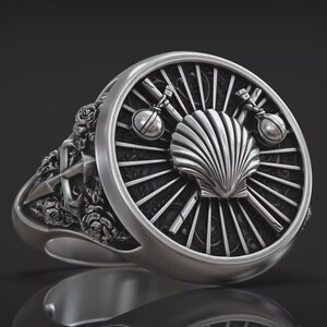 Può includere: Un anello d'argento dal design dettagliato. L'anello presenta una conchiglia centrale, affiancata da oggetti incrociati ed elementi circolari. La fascia è ornata da motivi floreali e nautici, creando un aspetto dettagliato e ornato.