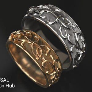 Könnte beinhalten: Zwei verzierte Ringe, einer goldfarben und einer silberfarben, mit floralem Design. Die Ringe liegen nebeneinander und zeigen die komplizierten Details der eingravierten Muster. Wahrscheinlich für eine Hochzeit oder Verlobung.