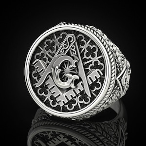 May include: A silver-colored ring with a detailed design. The ring features a central emblem with a compass, square, and the letter "G". The band has an intricate pattern. The ring is displayed against a black background.