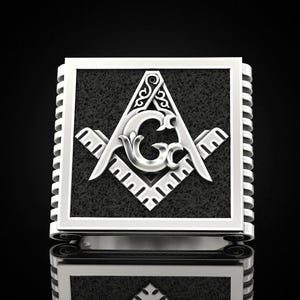 May include: Silver-colored ring featuring a square face with a textured black background. The ring showcases a Masonic symbol with a compass, square, and the letter "G". The ring has a detailed, ornate design.