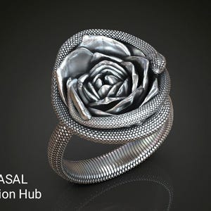 以下が含まれることがあります： バラとヘビのデザインが施されたシルバーリング。 リングはテクスチャ加工された表面です。 下部には「RASAL Fashion Hub」の文字が見えます。