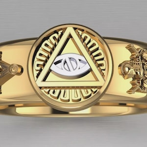 May include: A gold-colored ring with intricate Masonic symbols. The ring features a central design of a triangle with an eye, flanked by a square and compass on one side and a detailed emblem on the other. The ring has a polished finish.