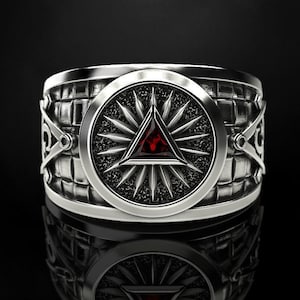 May include: A silver ring with a central circular design featuring a red triangle and starburst pattern. The ring has a textured band with geometric and decorative elements. The ring is displayed on a reflective black surface.