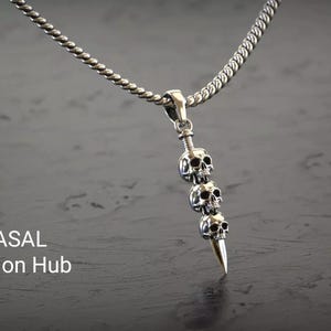 Può includere: Collana color argento con un ciondolo con tre ciondoli a forma di teschio e una punta. La catena ha un design a corda ritorta. Il testo "RASAL Fashion Hub" è visibile nell'immagine.