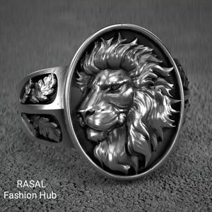 Könnte beinhalten: Ein silberner Ring mit einem detaillierten Löwenkopf in einem ovalen Rahmen. Das Ringband hat blattartige Designs an den Seiten. Der Text "RASAL Fashion Hub" ist unten sichtbar. Ein auffälliges Schmuckstück.