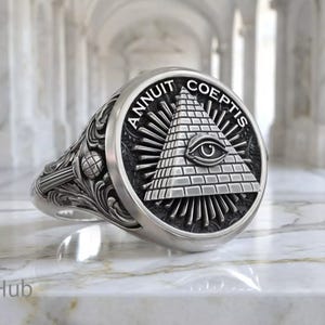 May include: Silver ring with an engraved design featuring the Eye of Providence within a pyramid, surrounded by the text "ANNUIT COEPTIS". The ring has ornate detailing on the band and is displayed on a marble surface.