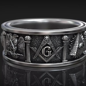 May include: A silver-colored ring with detailed Masonic symbols, including an eagle, compass and square, and an all-seeing eye. The ring has a textured band and is displayed against a dark background. The text "RASAL Fashion Hub" is visible.
