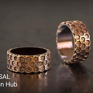 May include: Two bronze-colored rings with a honeycomb pattern. The rings are displayed on a dark surface. The text "RASAL Fashion Hub" is visible at the bottom of the image.