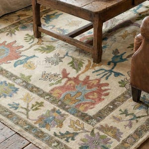 Puede incluir: Una alfombra beige con un estampado floral en tonos rojos, azules, verdes y amarillos. La alfombra está colocada debajo de una mesa de centro de madera y cerca de un sofá beige y un sillón de cuero. La alfombra tiene un borde.