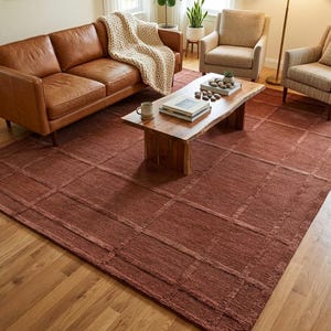 May include: A living room scene featuring a brown leather sofa, two armchairs, and a wooden coffee table. A large, rectangular, reddish-brown rug with a grid pattern covers the floor. Decorative items include a floor lamp, plants, and framed artwork.