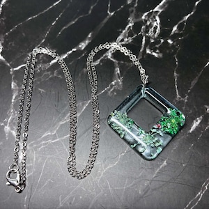 May include: A silver chain necklace with a square pendant. The pendant is clear with a diamond-shaped opening and contains green and red plant life. The necklace is displayed on a black marble surface.