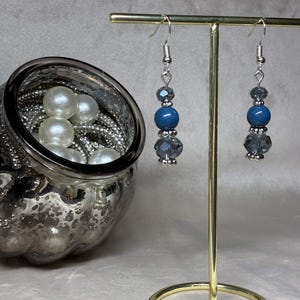 Pode incluir: Um par de brincos pendentes com uma conta azul central ladeada por contas cinzentas facetadas mais pequenas e espaçadores prateados, suspensos por ganchos prateados. Os brincos estão num suporte de metal dourado ao lado de um frasco de vidro.