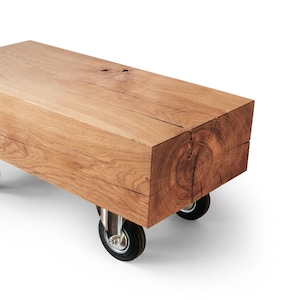 May include: A rectangular wooden coffee table made from a single block of light brown wood. The table rests on four small black and silver caster wheels, allowing for easy movement. The wood grain is visible.