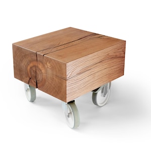 Puede incluir: Una mesa auxiliar rectangular de madera sobre ruedas. La mesa está hecha de un bloque macizo de madera, con vetas y grietas visibles. Las ruedas son grises y blancas. La madera tiene un color marrón cálido natural.