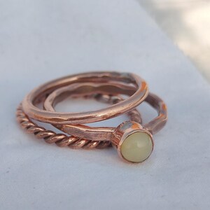 May include: A set of three copper rings. One ring has a twisted band, another has a smooth band, and the third features a pale yellow gemstone. The rings are stacked together on a white surface.