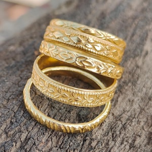 Op de afbeelding: Een stapel goudkleurige ringen met ingewikkelde bloemen- en textuurontwerpen. De ringen liggen op een ruw houten oppervlak, wat hun gedetailleerde vakmanschap laat zien. De ringen variëren in ontwerp, sommige met bladerpatronen en andere met getextureerde banden.