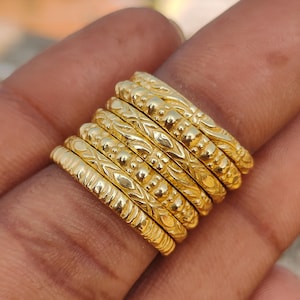 Puede incluir: Primer plano de siete anillos dorados apilados en un dedo. Cada anillo presenta un diseño texturizado único, con detalles de cuentas, grabados y patrones. La imagen destaca la artesanía y la variedad de diseños.