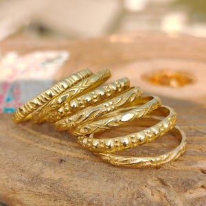 Puede incluir: Una colección de anillos dorados con varios patrones en relieve, incluyendo diseños de cuentas y florales. Los anillos están apilados sobre un trozo de madera flotante, mostrando sus intrincados detalles y cálidos tonos metálicos. Joyería.