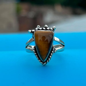 Op de afbeelding: Een zilveren ring met een driehoekige tijgeroog-edelsteen gezet in een decoratieve metalen zetting. De ringband heeft een gespleten ontwerp en de steen vertoont een goudbruine chatoyante glans met een glad, gepolijst oppervlak.