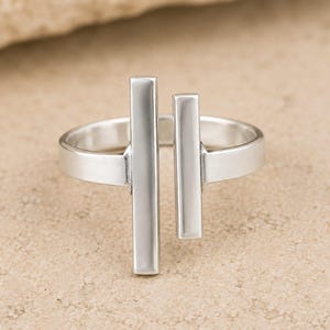 Puede incluir: Un anillo de tono plateado con un diseño de banda abierta que presenta dos barras rectangulares verticales de diferentes longitudes posicionadas paralelamente en el centro. El metal tiene un acabado liso y pulido que refleja la luz.
