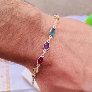 May include: A silver bracelet featuring a series of oval-shaped gemstones in various colors, including red, purple, blue, and yellow. The bracelet is worn on a wrist, showcasing the gemstones' vibrant hues and the intricate design of the silver links.