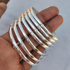May include: A set of seven polished silver bangles. The bangles are smooth, with a rounded profile, and reflect light. The jewelry is displayed on a light background. The brand name "KhushijewelCo" is visible.