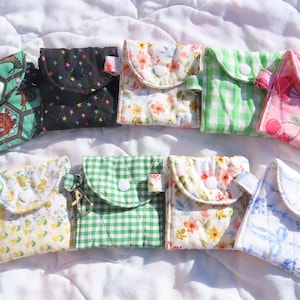 May include: A collection of small, fabric pouches in various patterns and colors, including floral, gingham, and star designs. Each pouch has a button closure and some have keychains attached. The pouches are arranged on a white, textured surface.