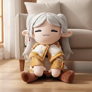 Puede incluir: Muñeca de peluche con expresión serena, cabello gris en coletas y atuendo blanco y dorado. La muñeca está sentada en un suelo de madera, con un sofá beige al fondo. Detalles rojos adornan las orejas de la muñeca.