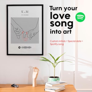 May include: A framed art print with a black border features a gray background with two hands intertwined, pinky fingers linked. The print includes initials "K & M" and the date "12.10.2022". Below is a Spotify code. Text reads "Turn your love song into art".
