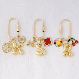 May include: Gold-colored keychains with various charms. One keychain has the letter "K" with a daisy and an oval charm. Another has the letter "S" with a balloon dog and a red flower. The last has the letter "R" with a flower and cherries.