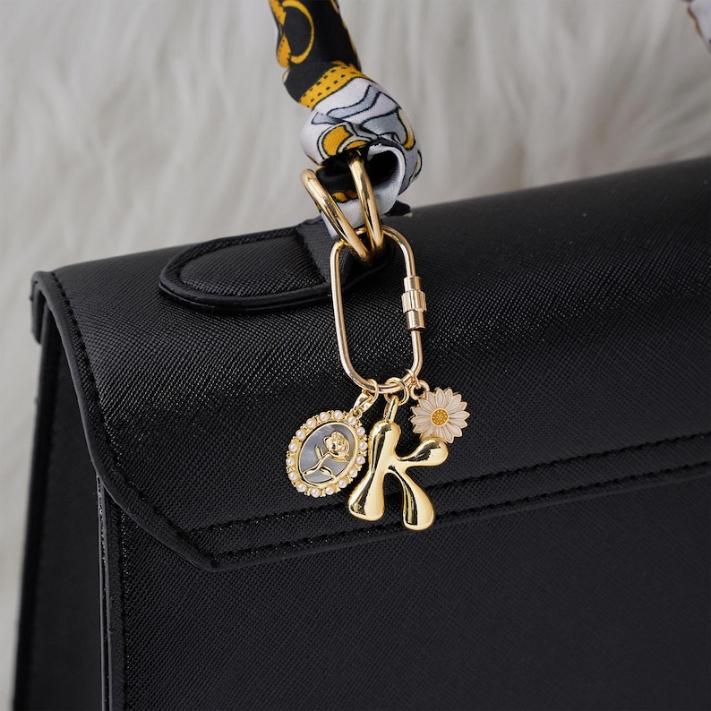 May include: A black handbag with a gold-colored charm. The charm includes a daisy, the letter "K", and an oval pendant with a rose design. A patterned scarf is attached to the handbag's handle.