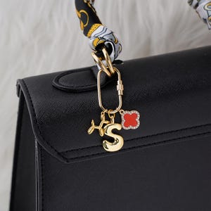 May include: A black handbag with a gold-colored charm. The charm includes the letter "S", a red clover, and a small airplane. The bag's handle is wrapped with a patterned scarf.