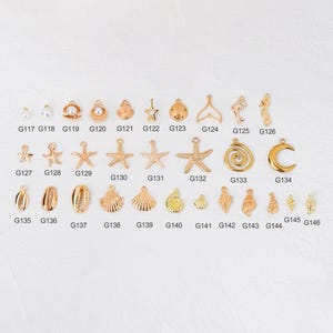 May include: A collection of gold-colored charms in various shapes, including seashells, starfish, pearls, and a crescent moon. The charms are arranged on a white surface and labeled with individual codes.